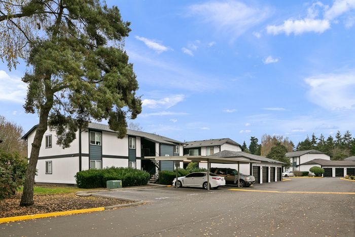 Charming apartment complex with modern design, surrounded by lush greenery and ample parking space, under a clear blue sky.
