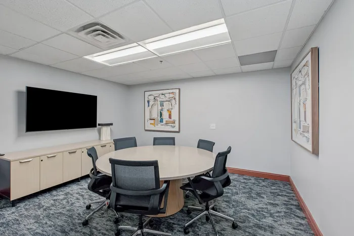 Modern conference room with a round table, six chairs, a large wall-mounted screen, and contemporary art on the wall.