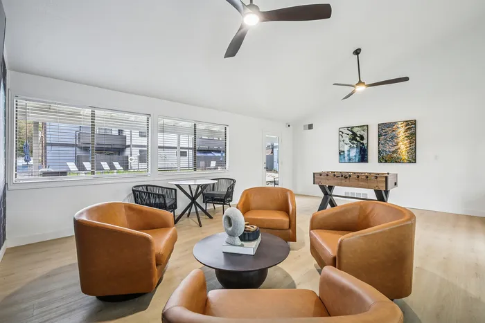 Modern apartment lounge with cozy leather chairs, round coffee table, and foosball table, featuring bright natural light through large windows.