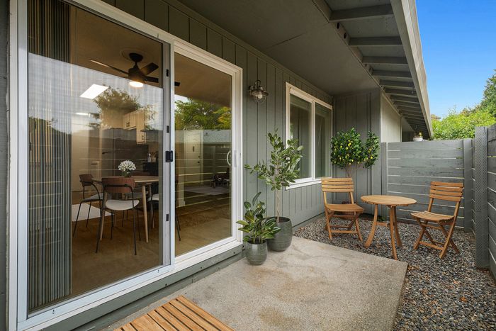 Charming outdoor patio with wooden furniture, lush greenery, and a view into a cozy dining area through sliding glass doors.