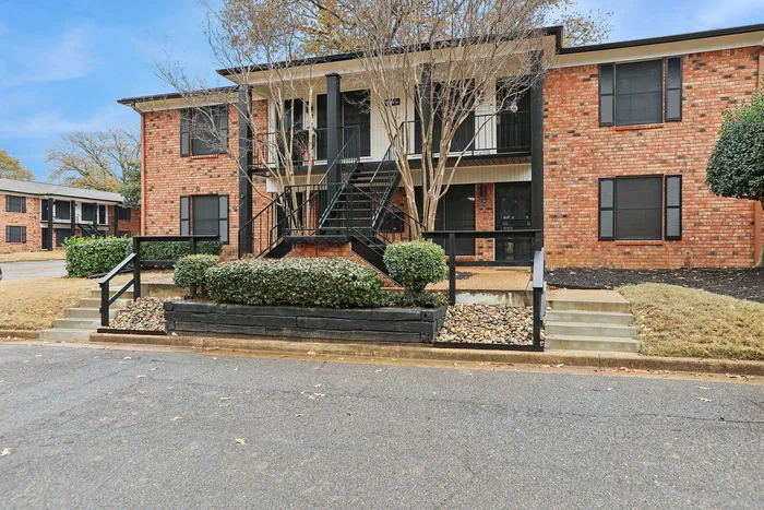 Charming brick apartment building with modern black accents, surrounded by lush greenery and elegant entrance steps.