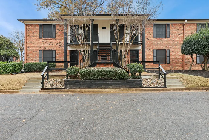 Charming brick apartment building with black shutters and stairway, surrounded by lush green landscaping and mature trees.