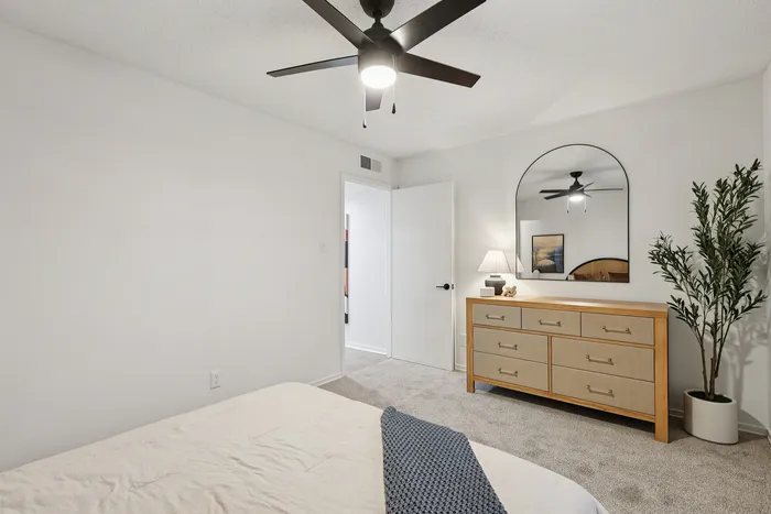 Contemporary bedroom with cozy, neutral tones featuring a sleek ceiling fan, elegant wooden dresser with mirror, and lush green plant.