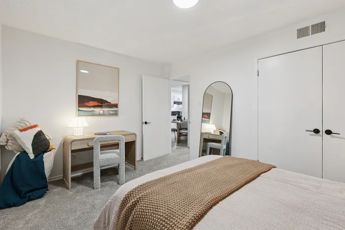 Inviting bedroom with modern decor, featuring a cozy bed, chic desk, elegant mirror, and soft carpet flooring.