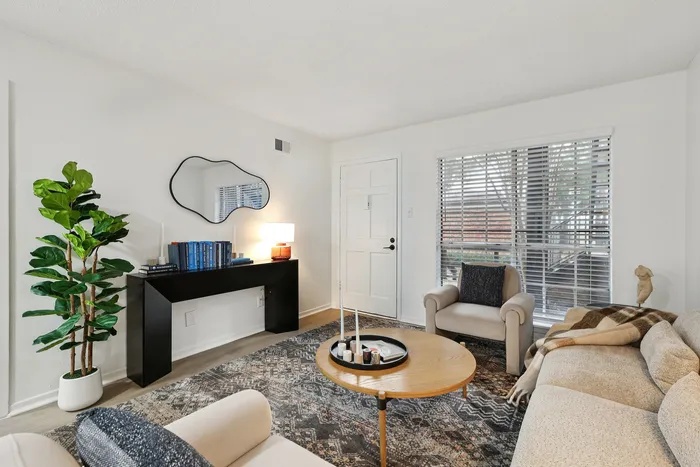 Bright and inviting living room featuring modern decor with a plush sofa, stylish armchair, and a chic coffee table. A sleek console with books and a contemporary mirror complements the space, enhanced by natural light through large windows and cozy ambiance from a warm-toned lamp.