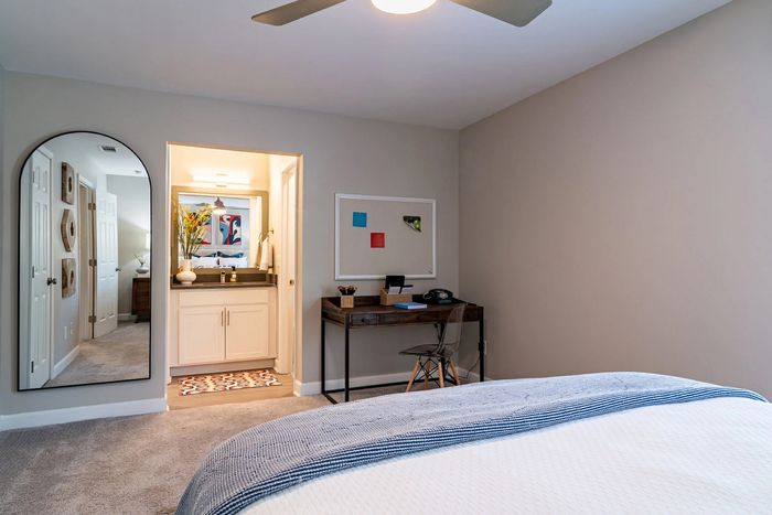 Cozy bedroom with modern decor, featuring a workspace, large mirror, and en-suite bathroom, illuminated by stylish overhead lighting.