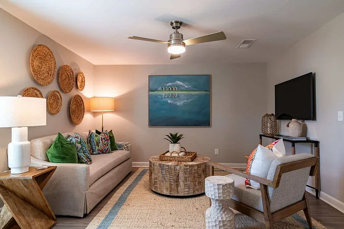 Chic living room with cozy beige sofa, vibrant patterned cushions, and rustic wood accents. Modern ceiling fan, artistic wall decor, and inviting lighting create a warm ambiance.