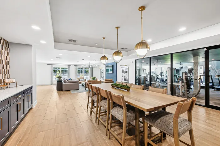 Expansive open-concept living area showcasing a stylish dining setup with pendant lighting, cozy seating, and a view into a modern fitness center.