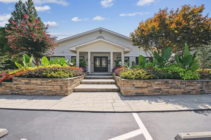 Charming apartment entrance with vibrant landscaping, blooming flowers, and elegant stonework under a sunny blue sky.