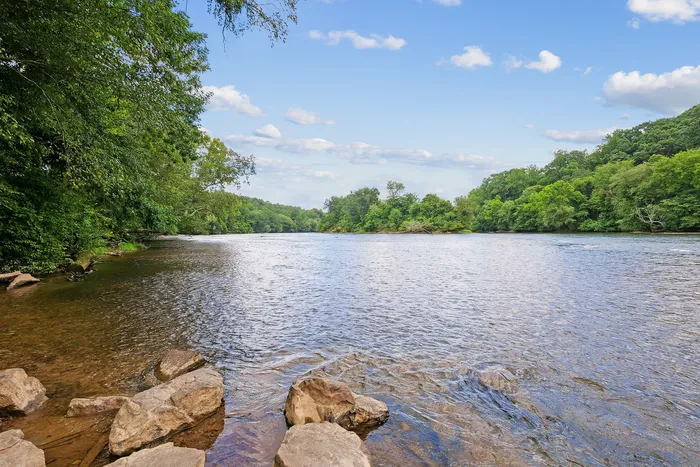 Tranquil riverside view with lush green trees and a serene blue sky, perfect for relaxation and nature enthusiasts.