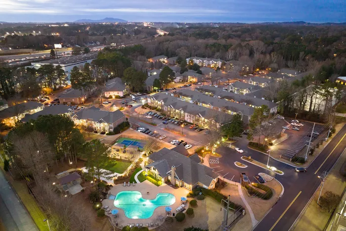 Scenic aerial view of a charming apartment complex with a sparkling pool, tennis courts, and lush greenery, nestled in a serene residential area.