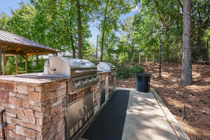 Inviting outdoor BBQ area with modern grills and a rustic stone counter, nestled among lush greenery and serene trees.