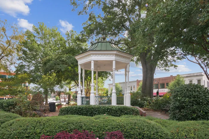 Charming white gazebo nestled in a lush green park, surrounded by vibrant trees and quaint town buildings under a bright blue sky.