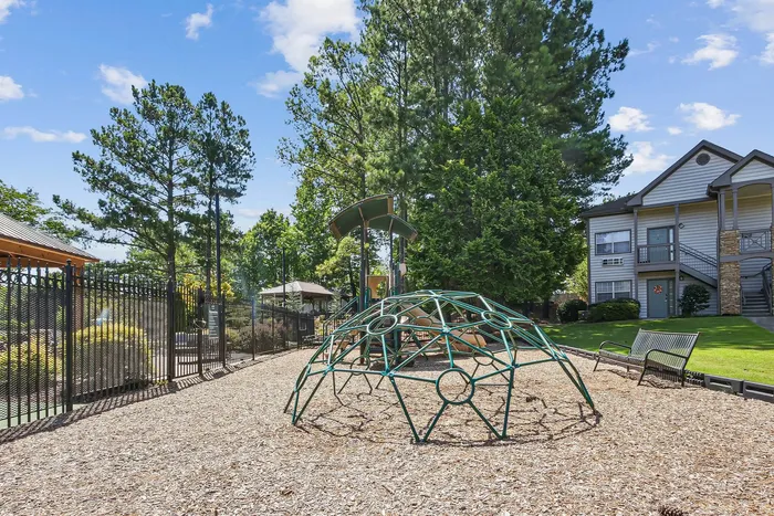Charming outdoor play area with a dome climber, set against a backdrop of lush greenery and cozy apartments, perfect for family-friendly living.