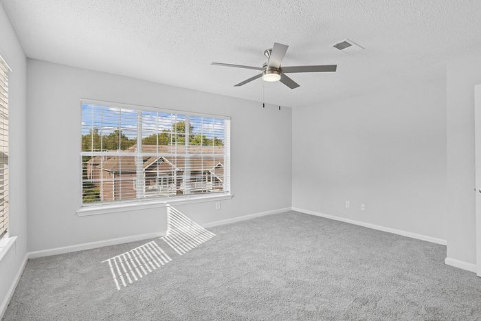 Bright and airy room with plush carpeting, large window view, and sleek ceiling fan, offering a modern and inviting atmosphere.