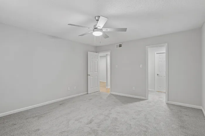 Bright, spacious bedroom featuring soft carpet flooring, a modern ceiling fan, and freshly painted walls with two doors leading to adjacent rooms, perfect for comfortable living.