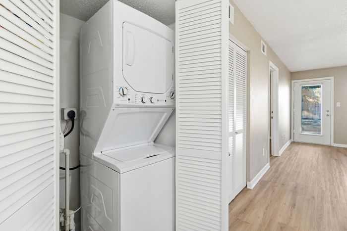 Townhomes at TSC Bright hallway featuring a modern in-unit laundry with stackable washer and dryer, sleek hardwood floors, and access to a private outdoor area through a glass door.