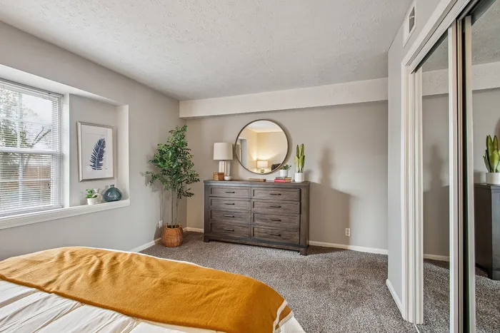 Inviting bedroom with soft carpet, a cozy bed adorned with a warm mustard throw, and elegant decor featuring a dark wooden dresser with a round mirror reflecting natural light from a large window.