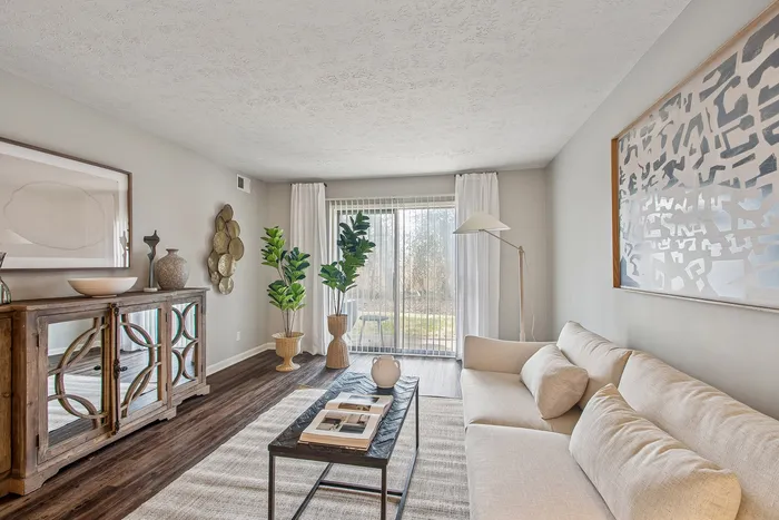 Bright and inviting living room with a stylish beige sofa, modern art, and natural light streaming through large windows, complemented by elegant decor and lush indoor plants.
