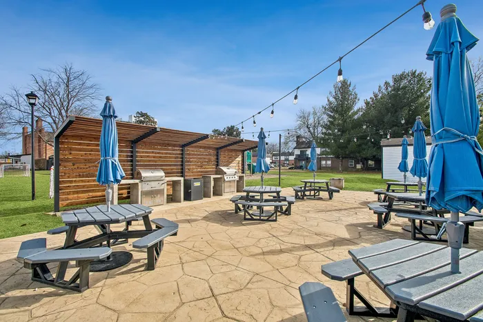 Inviting outdoor common area featuring picnic tables with blue umbrellas, string lights, and modern grilling stations set against a sunny backdrop.