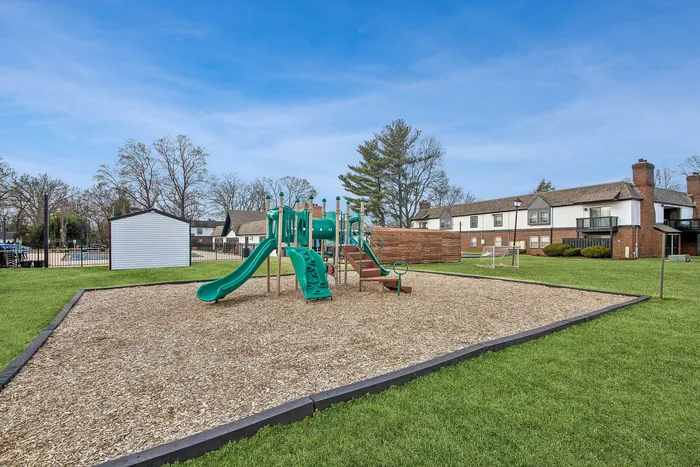 Charming outdoor playground with green slides and climbing structures, nestled amidst a scenic residential area with manicured lawns and townhouses.