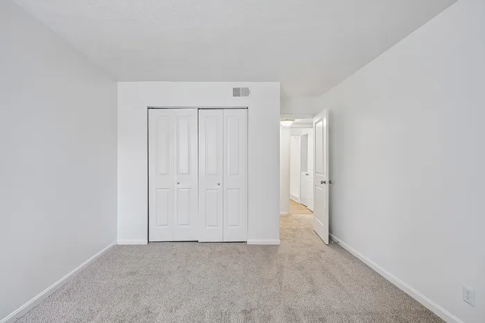 Bright and inviting empty room with plush carpeting, featuring a double-door closet and an open door leading to a hallway, perfect for a customizable living space.