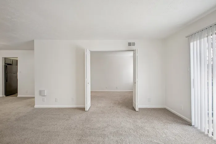 Bright, open-concept apartment with plush carpeting and a spacious entryway leading to a separate room, accented by large windows for natural light.