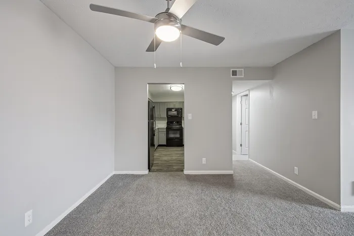 Modern apartment interior with plush carpeted floors and a sleek ceiling fan, leading to a stylish kitchen with contemporary appliances.