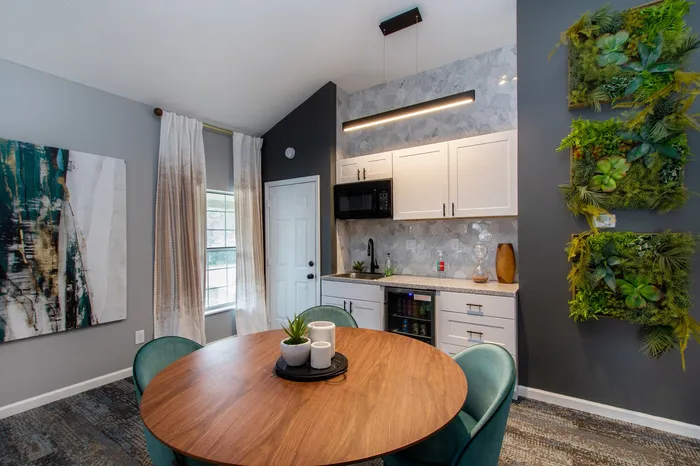 Modern dining area featuring a stylish round wooden table, elegant teal chairs, vibrant wall art, and a sleek kitchenette with white cabinets and lush vertical greenery.