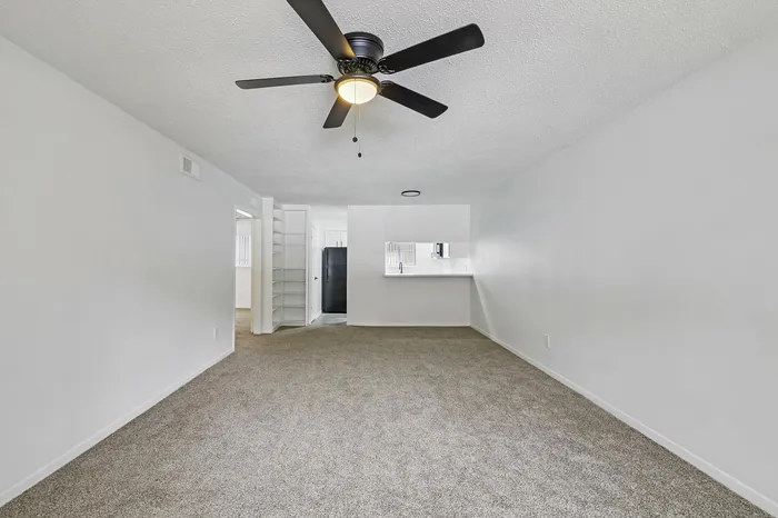 Bright and spacious living room with plush carpet, modern ceiling fan, and open layout leading to kitchen area.