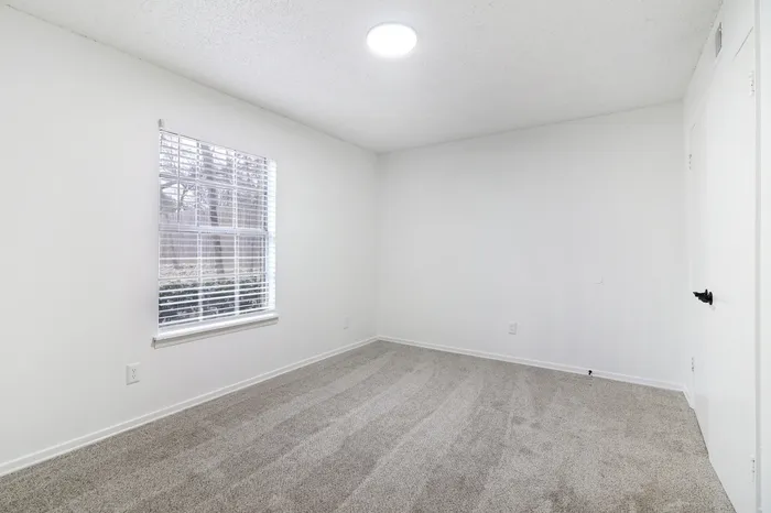 Bright and spacious bedroom with plush carpeting, large window allowing natural light, and fresh white walls perfect for personalization.