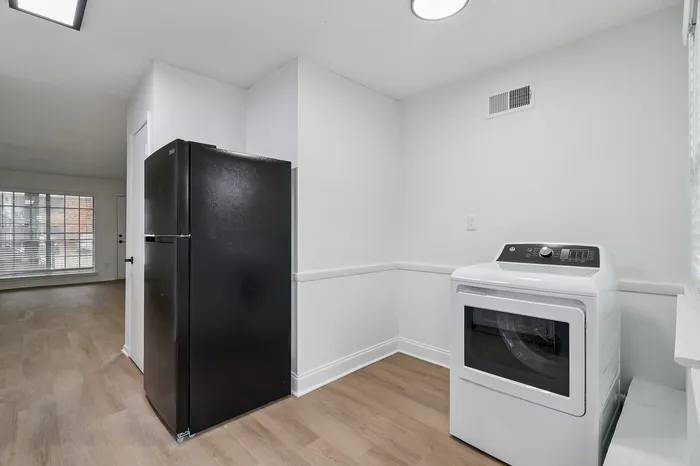 Modern laundry area featuring sleek black refrigerator and state-of-the-art white washer against a backdrop of elegant wood flooring and bright window lighting.