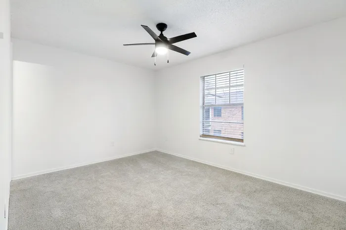 Bright and airy room with plush carpet, large window, and modern ceiling fan, ideal for a cozy bedroom or inviting office space.