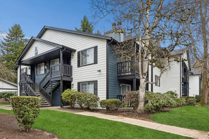 Charming two-story apartment complex with white siding, surrounded by lush greenery and manicured lawns, featuring outdoor stairs and a serene neighborhood atmosphere.