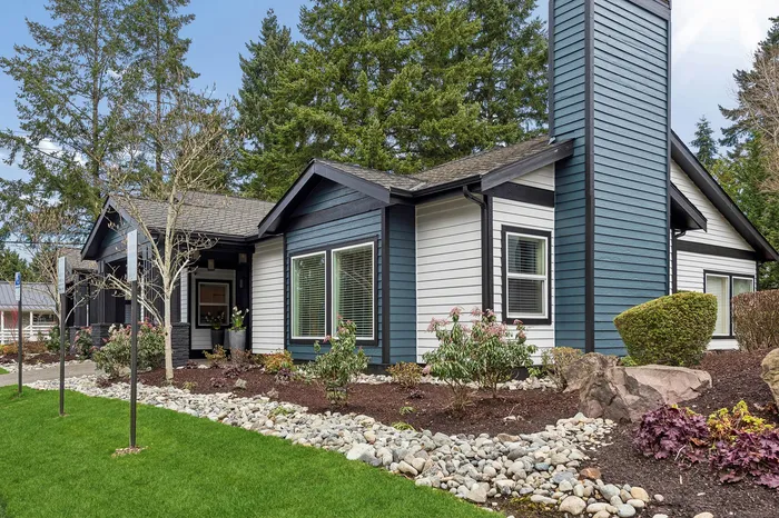Charming modern home with elegant blue and white facade, lush landscaping, and scenic backdrop of tall trees.