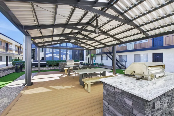 Modern outdoor pavilion with sleek barbecue area and seating, surrounded by lush greenery and contemporary apartment architecture.