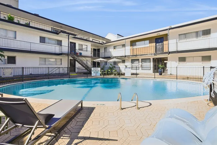 Bright and inviting apartment complex featuring a sparkling outdoor pool with lounge chairs, perfect for relaxation under the sunny sky.