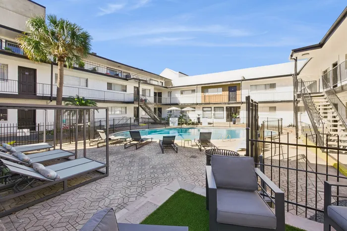 Inviting apartment courtyard featuring a sparkling pool, stylish lounge chairs, and tropical landscaping under a brilliant blue sky.