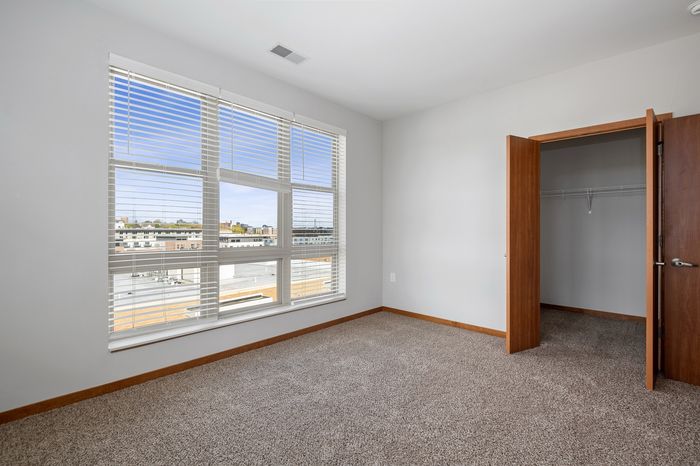 Bright and airy bedroom with large windows offering a stunning cityscape view, featuring a spacious closet with wooden doors and plush carpet flooring.