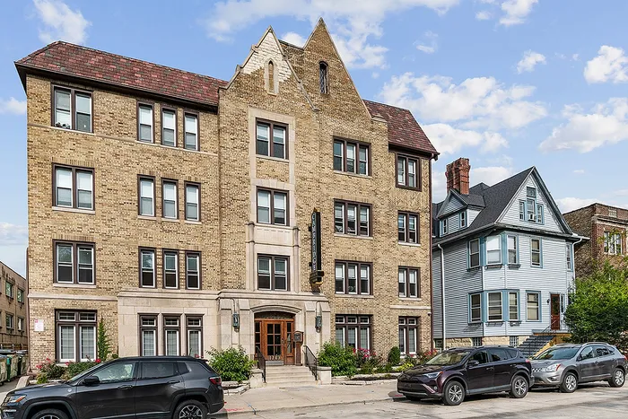 Charming brick apartment building with classic architecture, surrounded by lush greenery and parked cars under a bright blue sky.