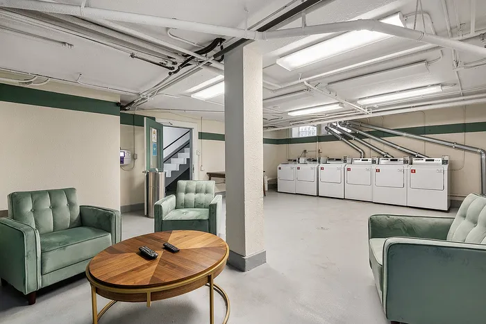 Inviting modern laundry room with comfortable seating area, featuring plush green armchairs and a stylish wooden table, ideal for relaxation while doing laundry.