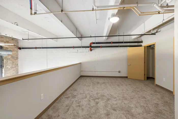 Modern loft space with exposed brick and piping, featuring a cozy carpeted area and an inviting open doorway.