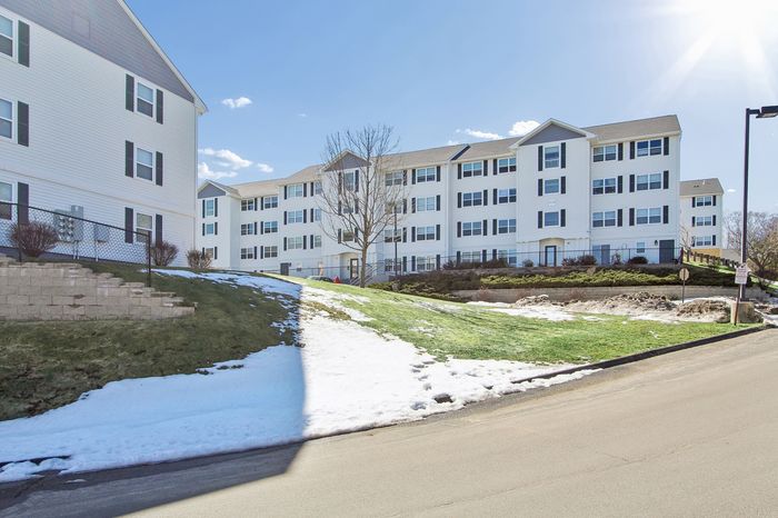 Modern four-story apartment building with charming dormer windows, surrounded by lush greenery and a light dusting of snow, under a bright blue sky.