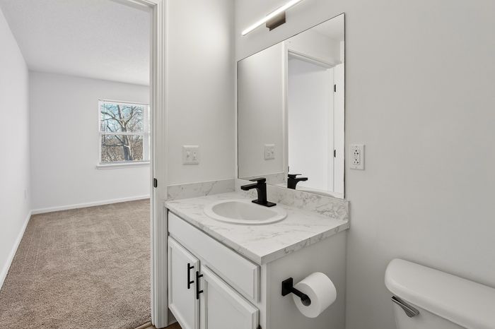 Modern bathroom with sleek design, featuring a stylish vanity and large mirror, adjacent to a bright, carpeted bedroom with natural light.