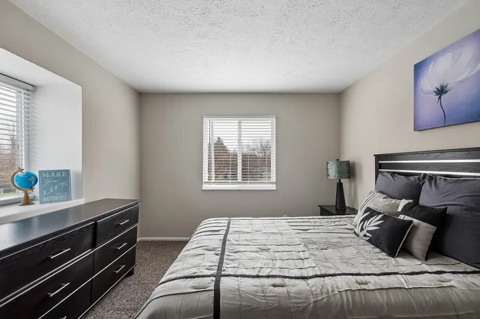 Bright and stylish bedroom featuring a plush bed, modern dresser, and serene decor with natural light streaming through large windows.