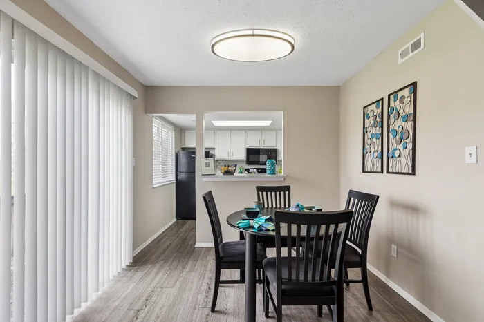 Bright dining area with elegant lighting, modern table, and a sleek view into a stylish kitchen. Natural light filters through large vertical blinds.