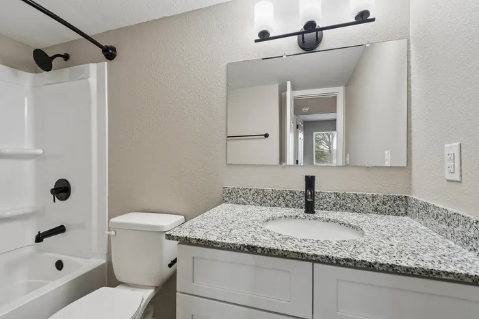 Modern bathroom featuring a sleek granite countertop, elegant lighting over a large mirror, and a pristine shower-tub combination.