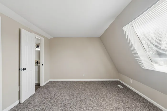Bright, spacious bedroom with plush carpet, angled ceiling, and large window with blinds, perfect for relaxation and comfort.