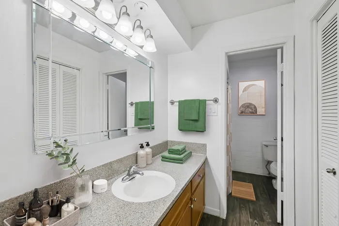 Elegant bathroom with granite countertops, a large mirror with modern lighting, and green towel accents.