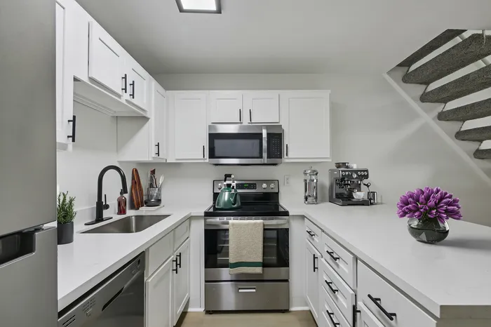 Modern white kitchen with sleek cabinetry, stainless steel appliances, and a stylish coffee machine, enhanced by a pop of color from a vase of purple flowers.